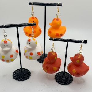 May include: Four pairs of rubber duck earrings with fall leaf designs. The ducks are orange, white, and red with orange and yellow leaves. The earrings are on black earring stands.