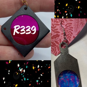 May include: A pair of earrings with a black and silver metal base. One earring has a pink and red circle with the text "R339" inside. The other earring has a blue and purple circle with a swirling pattern inside.