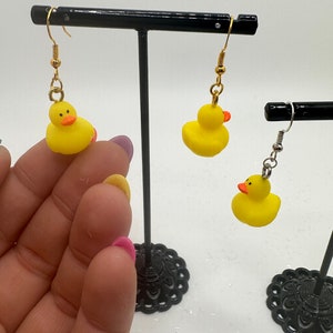 Könnte beinhalten: Ein Paar gelbe Gummiente-Ohrringe mit goldenen Haken. Die Ohrringe werden auf einem schwarzen Ohrringständer ausgestellt.