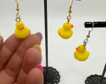 May include: A pair of yellow rubber ducky earrings with gold hooks. The earrings are displayed on a black earring stand.