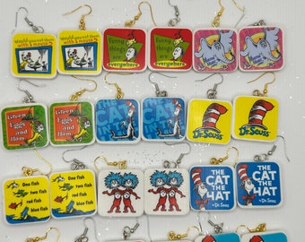 Puede incluir: Un conjunto de 18 pares de pendientes con personajes y escenas del libro de Dr. Seuss, El Gato en el Sombrero. Los pendientes están hechos de madera y tienen forma cuadrada. Algunos de los pendientes presentan al Gato en el Sombrero, Cosa 1 y Cosa 2, y el Lorax.