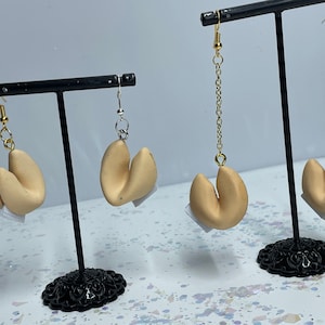 Pode incluir: Um par de brincos de biscoito da sorte. Os brincos são feitos de argila polimérica e têm o formato de biscoitos da sorte. Os brincos estão em um suporte de brincos preto. Os brincos estão disponíveis em dois estilos: um com uma corrente dourada e outro com uma corrente prateada.