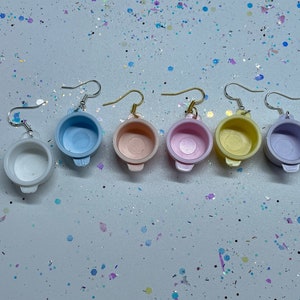 Può includere: Sei paia di orecchini a forma di pentola di colore pastello. Gli orecchini sono in plastica e hanno un gancio argentato. I colori degli orecchini sono bianco, azzurro, rosa, giallo e lavanda.