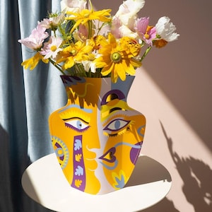 May include: A yellow ceramic vase with a colorful abstract face design. The vase is filled with yellow, white, and pink flowers.