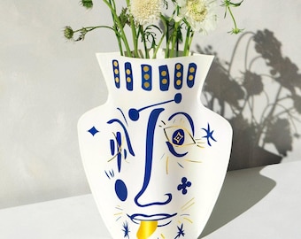 May include: White ceramic vase with a blue and gold face design. The vase has a wide mouth and a narrow base. The face has a blue eye, a gold tongue, and a gold star on the cheek. The vase is filled with white flowers.