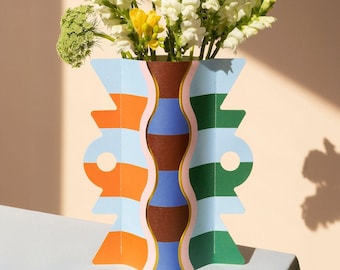 May include: A colorful, geometric vase with a blue, orange, green, and brown striped pattern. The vase is made of paper and has a unique, folded design. The vase is filled with white and yellow flowers.
