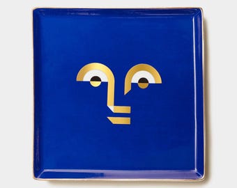May include: A square blue ceramic tray with a gold rim and a gold geometric face design.