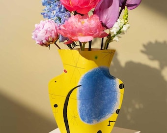 May include: A yellow ceramic vase with a blue abstract design and black accents holds a vibrant bouquet of pink peonies, blue delphiniums, and purple anthuriums. The vase sits on a white surface against a light yellow background.