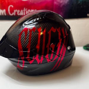 Puede incluir: Un casco de motocicleta de fibra de carbono negra con acabado brillante y letras rojas grandes y estilizadas en el lateral. El casco incluye una visera oscura, puertos de ventilaci&oacute;n en la parte superior y un detalle de tornillo plateado.