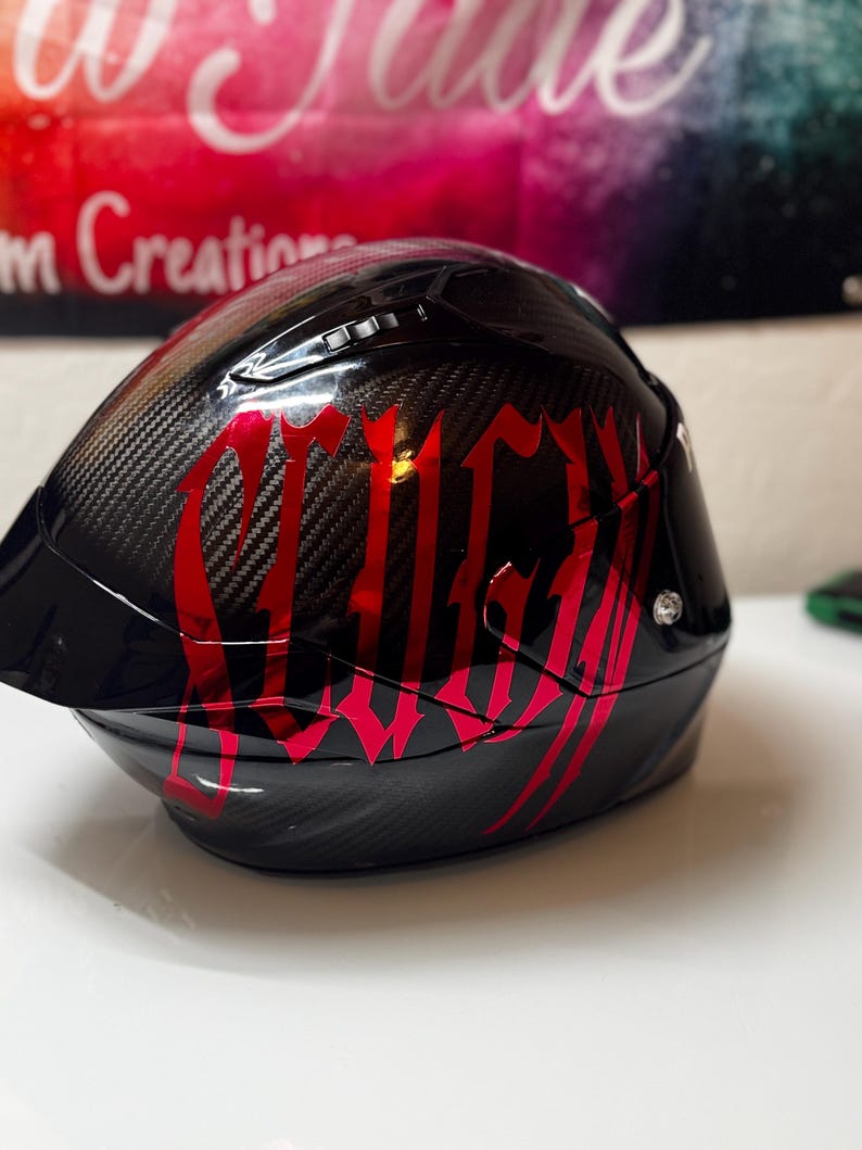 Puede incluir: Un casco de motocicleta de fibra de carbono negra con acabado brillante y letras rojas grandes y estilizadas en el lateral. El casco incluye una visera oscura, puertos de ventilaci&oacute;n en la parte superior y un detalle de tornillo plateado.