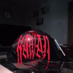 Puede incluir: Un casco de motocicleta negro con textura de fibra de carbono y letras rojas grandes y estilizadas que dicen KAWAI. El casco cuenta con una visera tintada oscura e incluye el texto @kawaii_degenerate impreso verticalmente en el lateral.