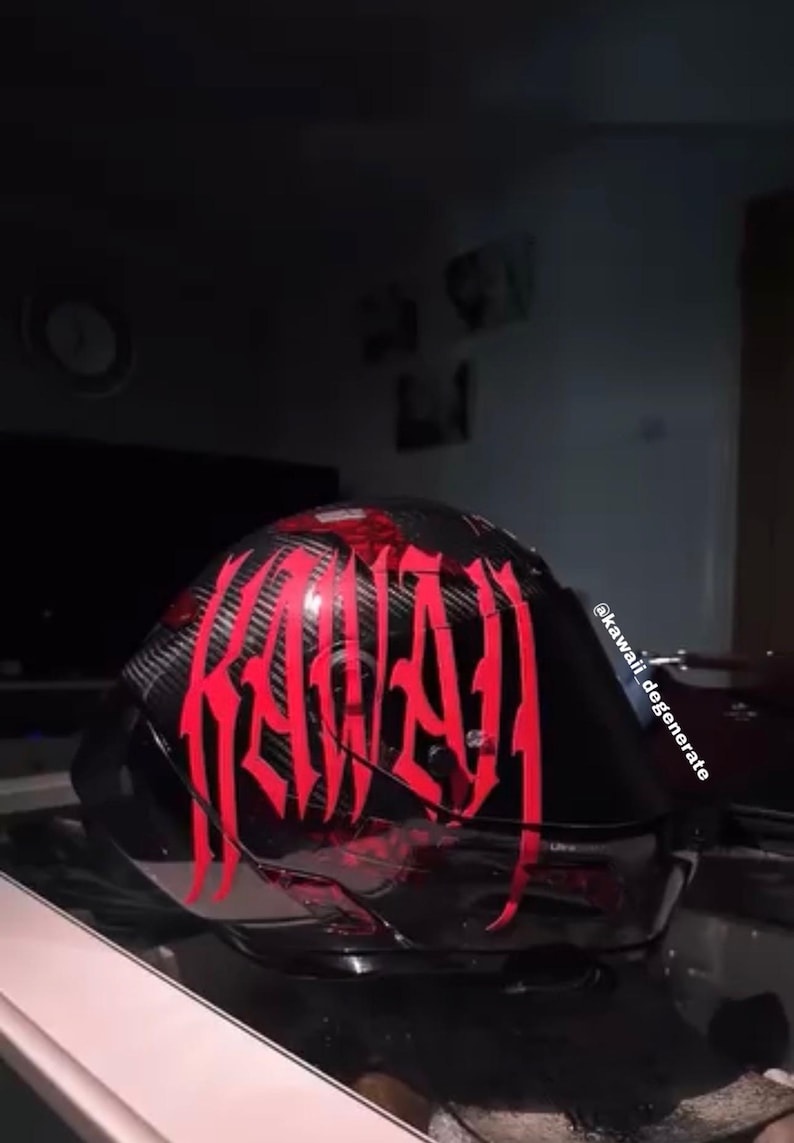 Puede incluir: Un casco de motocicleta negro con textura de fibra de carbono y letras rojas grandes y estilizadas que dicen KAWAI. El casco cuenta con una visera tintada oscura e incluye el texto @kawaii_degenerate impreso verticalmente en el lateral.