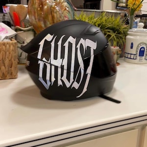Puede incluir: Un casco de motocicleta negro mate con la palabra GHOST escrita en letras plateadas reflectantes, grandes y estilizadas en el lateral. El casco incluye una visera tintada oscura y un dise&ntilde;o de carcasa aerodin&aacute;mica lisa con correa visible.