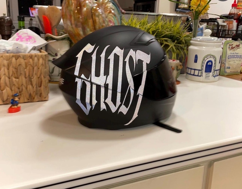 Puede incluir: Un casco de motocicleta negro mate con la palabra GHOST escrita en letras plateadas reflectantes, grandes y estilizadas en el lateral. El casco incluye una visera tintada oscura y un dise&ntilde;o de carcasa aerodin&aacute;mica lisa con correa visible.