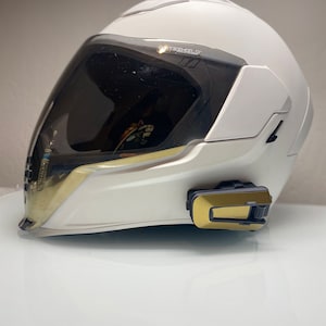 May include: A white motorcycle helmet with a tinted visor and gold accents. The helmet has a sleek design and a black communication device attached to the side. The visor has the text "TEXSHIELD".