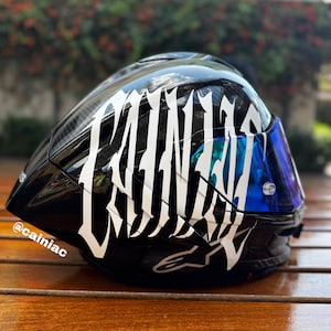 Puede incluir: Un casco de motocicleta negro brillante con grandes letras blancas estilizadas que forman la palabra CAINIAC en el lateral. El casco incluye una visera tintada en azul reflectante y el texto @cainiac impreso en letras blancas peque&ntilde;as en la base.