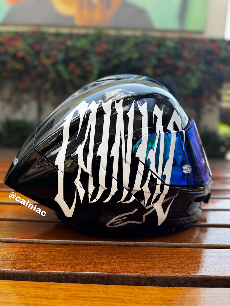 Puede incluir: Un casco de motocicleta negro brillante con grandes letras blancas estilizadas que forman la palabra CAINIAC en el lateral. El casco incluye una visera tintada en azul reflectante y el texto @cainiac impreso en letras blancas peque&ntilde;as en la base.