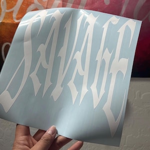 Puede incluir: Una calcoman&iacute;a de vinilo blanca con la palabra SAVAGE escrita en una fuente g&oacute;tica estilizada de bordes afilados. La calcoman&iacute;a se presenta sobre una hoja de soporte azul claro, mostrando el intrincado dise&ntilde;o de letras puntiagudas.