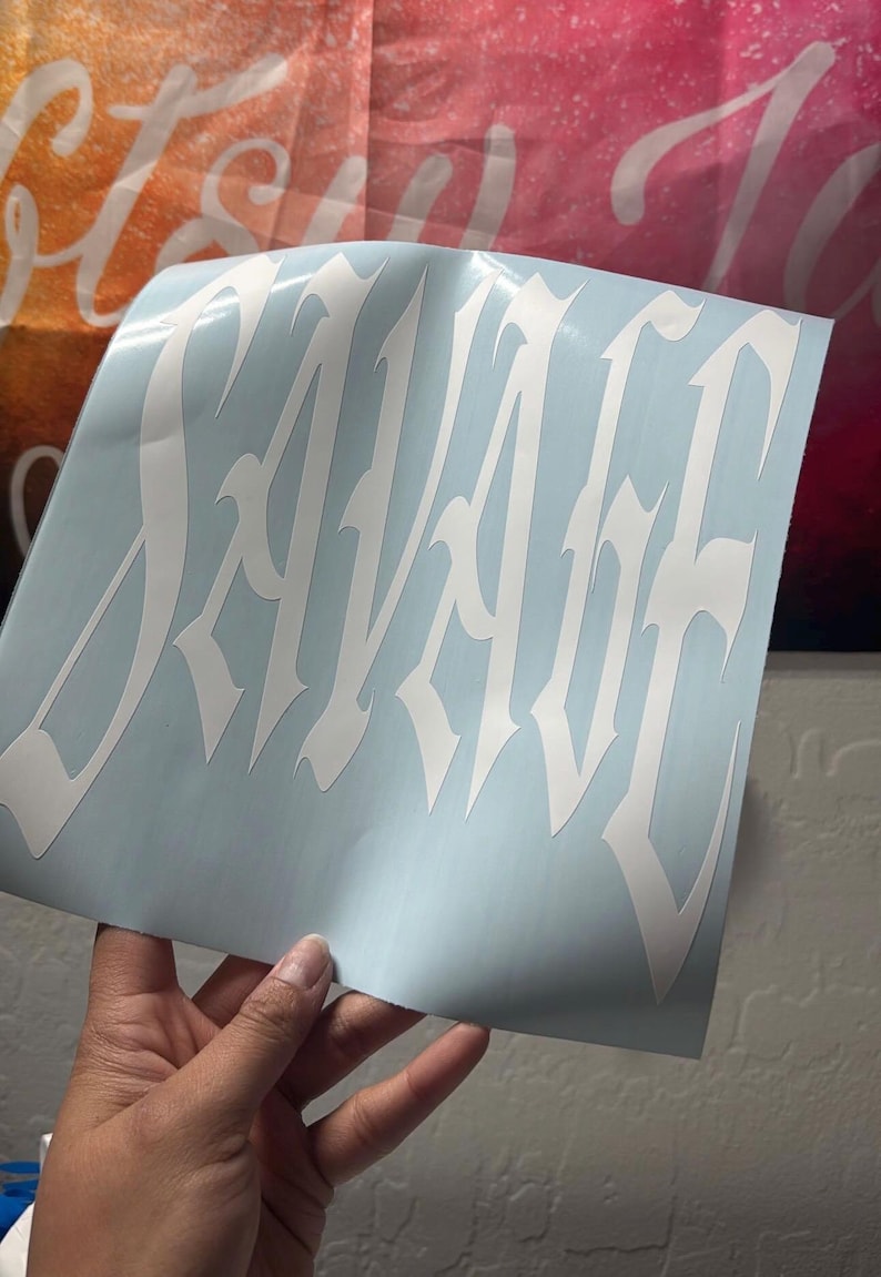 Puede incluir: Una calcoman&iacute;a de vinilo blanca con la palabra SAVAGE escrita en una fuente g&oacute;tica estilizada de bordes afilados. La calcoman&iacute;a se presenta sobre una hoja de soporte azul claro, mostrando el intrincado dise&ntilde;o de letras puntiagudas.