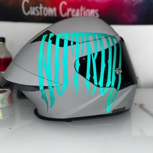 Puede incluir: Un casco de moto integral de color gris mate con un gran adhesivo gr&aacute;fico de color verde azulado brillante en el lateral. El casco incluye una visera tintada oscura y un borde negro a lo largo de la parte inferior, con un dise&ntilde;o aerodin&aacute;mico.