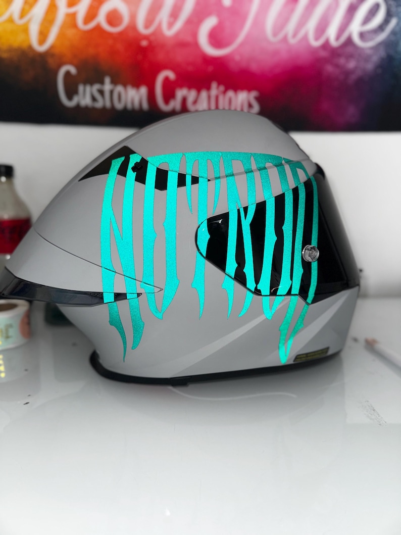 Puede incluir: Un casco de moto integral de color gris mate con un gran adhesivo gr&aacute;fico de color verde azulado brillante en el lateral. El casco incluye una visera tintada oscura y un borde negro a lo largo de la parte inferior, con un dise&ntilde;o aerodin&aacute;mico.