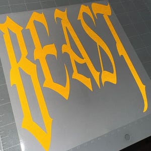 Puede incluir: Una calcoman&iacute;a de vinilo de color amarillo brillante con la palabra BEAST escrita en una fuente g&oacute;tica de &aacute;ngulos afilados. La calcoman&iacute;a est&aacute; sobre una hoja de soporte gris claro lisa que incluye una cuadr&iacute;cula y marcas num&eacute;ricas.