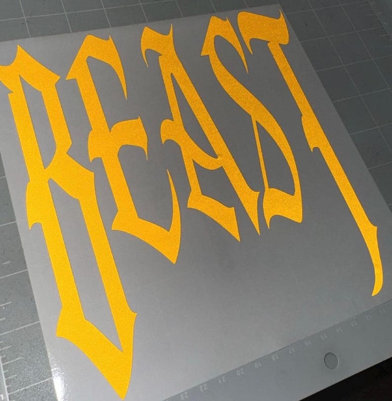 Puede incluir: Una calcoman&iacute;a de vinilo de color amarillo brillante con la palabra BEAST escrita en una fuente g&oacute;tica de &aacute;ngulos afilados. La calcoman&iacute;a est&aacute; sobre una hoja de soporte gris claro lisa que incluye una cuadr&iacute;cula y marcas num&eacute;ricas.