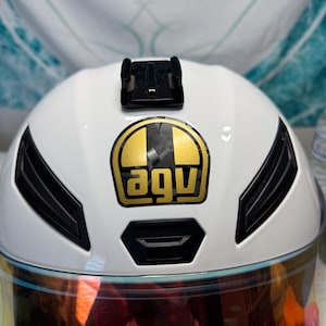 Puede incluir: Casco de moto blanco con detalles negros y un logotipo "agv" dorado y negro. El casco tiene una visera tintada y un soporte de montaje negro. Diseñado para la seguridad y la protección.