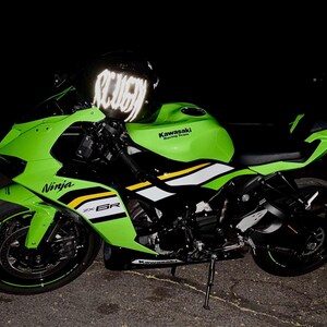 Puede incluir: Una motocicleta deportiva Kawasaki Ninja ZX-6R de color verde lima y negro con calcoman&iacute;as laterales blancas y amarillas, ruedas negras con cinta de borde verde y un casco negro con letras blancas reflectantes sobre el manillar de noche.