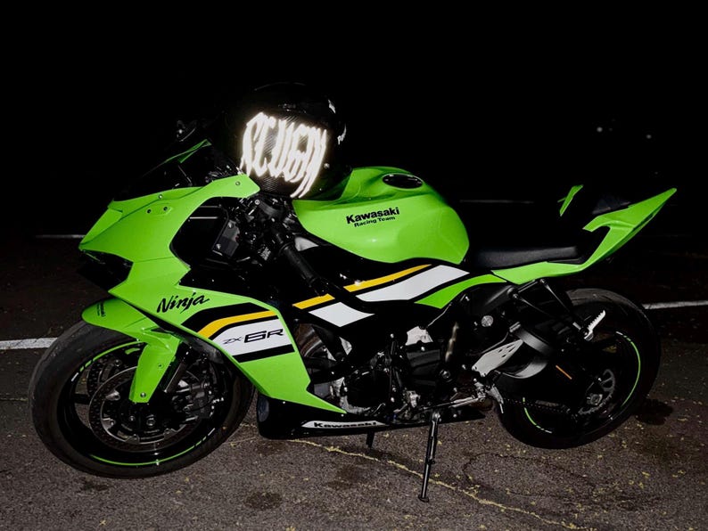 Puede incluir: Una motocicleta deportiva Kawasaki Ninja ZX-6R de color verde lima y negro con calcoman&iacute;as laterales blancas y amarillas, ruedas negras con cinta de borde verde y un casco negro con letras blancas reflectantes sobre el manillar de noche.