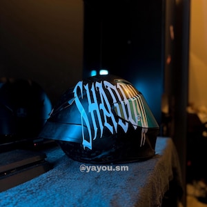 Puede incluir: Un casco de motocicleta negro con un texto gr&aacute;fico blanco grande y estilizado que dice SHADOW en el lateral. El casco tiene una visera oscura y reflectante e incluye el peque&ntilde;o texto blanco @yayou.sm impreso cerca del borde inferior.