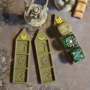 May include: A selection of miniature war game terrain pieces. The set includes several small, detailed artillery pieces, two green markers with arrow symbols, and a dice holder with the word "FIRE". The pieces are primarily grey and green.