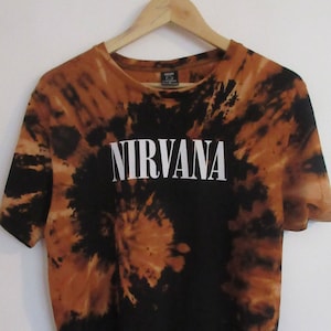 Może przedstawiać: Koszulka typu tie-dye z napisem "NIRVANA" w białych literach. Koszulka ma spiralny wzór w odcieniach czerni i brązu. Koszulka wisi na drewnianym wieszaku.