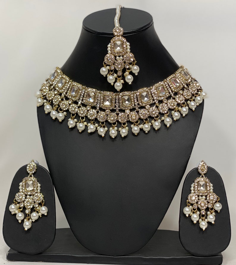 May include: A gold-toned bridal jewelry set displayed on a black mannequin. The set includes a necklace, earrings, and a headpiece, all adorned with clear square-cut stones and small white pearls. The necklace and earrings have a cascading design.