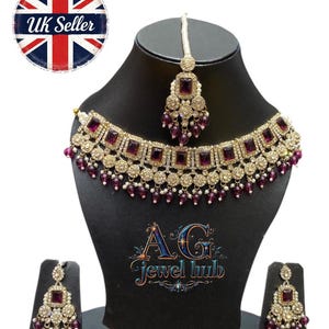 May include: A gold-toned necklace, earrings, and headpiece set with square, deep purple stones and small, dangling purple beads. The jewelry is displayed on a black mannequin head and stand. The set is from AG Jewel Hub, a UK seller.