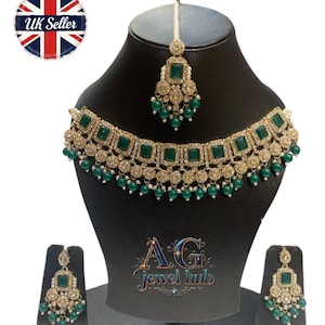 May include: A gold-toned jewelry set featuring emerald green square-cut stones and small green bead accents. The set includes a necklace, earrings, and a headpiece, all displayed on a black mannequin. The text "AG Jewel Hub" is visible.