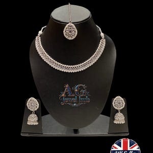 K&ouml;nnte beinhalten: Ein silberfarbenes Schmuckset, bestehend aus Halskette, Ohrringen und Kopfschmuck, pr&auml;sentiert auf einer schwarzen B&uuml;ste. Die Halskette und die Ohrringe weisen detaillierte Designs auf. Der Text "Exclusive New Design in Silver colour" ist sichtbar.