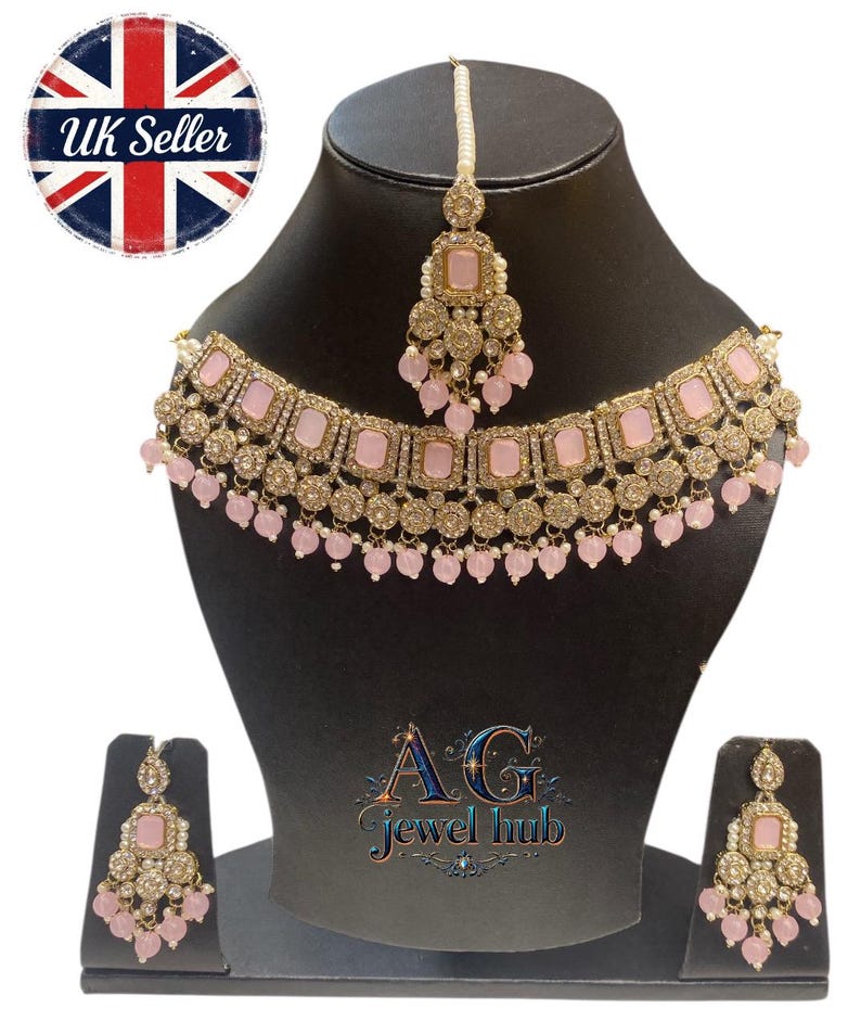 May include: A gold-toned jewelry set featuring a necklace, earrings, and a headpiece. The set includes pink square-shaped stones and small pink bead accents. The jewelry is displayed on a black mannequin head and stand. The text "AG Jewel hub" is visible.
