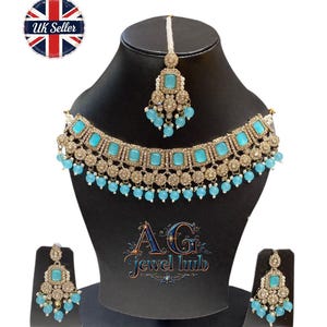 May include: A gold-toned jewelry set featuring a necklace, earrings, and a headpiece, all adorned with square turquoise-colored stones and small blue beads. The set is displayed on a black mannequin head. The logo "AG Jewel Hub" is visible.