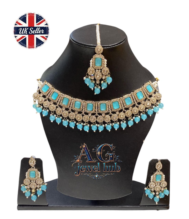 May include: A gold-toned jewelry set featuring a necklace, earrings, and a headpiece, all adorned with square turquoise-colored stones and small blue beads. The set is displayed on a black mannequin head. The logo "AG Jewel Hub" is visible.