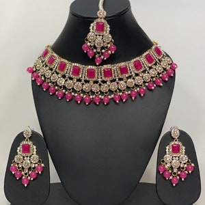 May include: A three-piece jewelry set featuring a necklace, earrings, and a headpiece. The set includes a gold-toned necklace with square magenta stones and clear crystals. The earrings and headpiece mirror the necklace design, with magenta beads and white pearls.