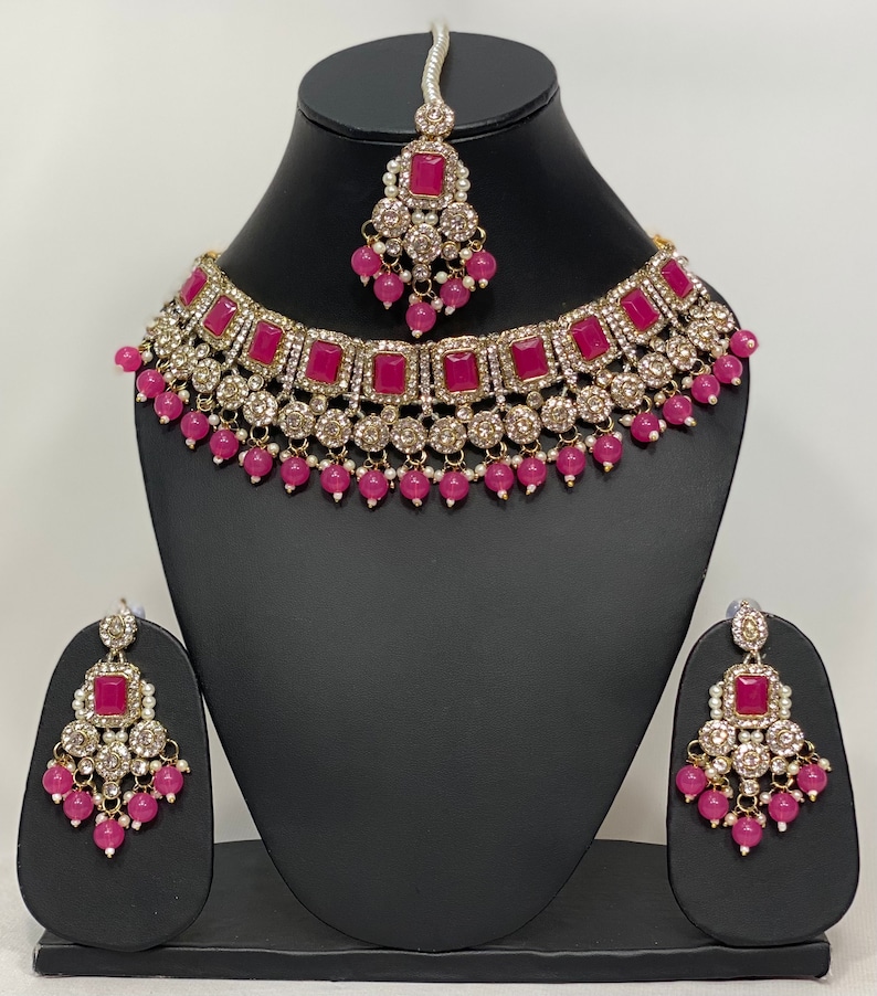 May include: A three-piece jewelry set featuring a necklace, earrings, and a headpiece. The set includes a gold-toned necklace with square magenta stones and clear crystals. The earrings and headpiece mirror the necklace design, with magenta beads and white pearls.