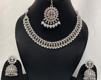 May include: A silver and white bridal jewelry set with a necklace, earrings, and a maang tikka. The necklace features a delicate design with small white pearls and rhinestones. The earrings are jhumka style with a round shape and a dangling pearl. The maang tikka is a traditional Indian headpiece with a round design and a chain.