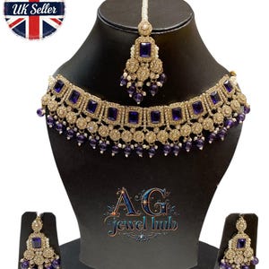 May include: A gold-toned jewelry set featuring a necklace, earrings, and a headpiece, all adorned with square, deep blue gemstones and small, dangling purple beads. The set is displayed on a black mannequin. The text "AG Jewel hub" is visible.
