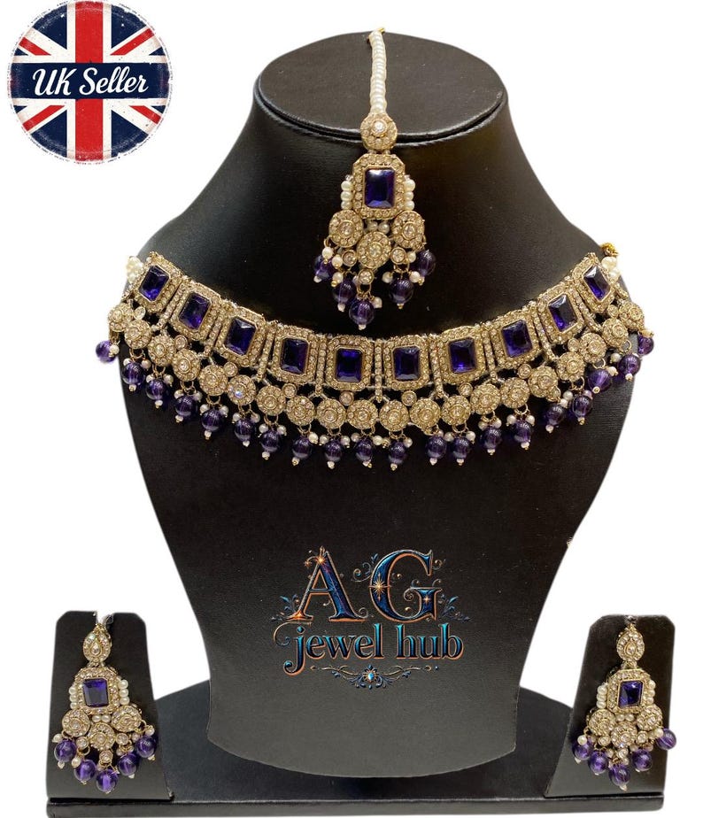 May include: A gold-toned jewelry set featuring a necklace, earrings, and a headpiece, all adorned with square, deep blue gemstones and small, dangling purple beads. The set is displayed on a black mannequin. The text "AG Jewel hub" is visible.