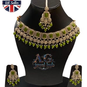 May include: An olive green and silver-toned jewelry set, including a necklace, earrings, and a headpiece. The necklace and headpiece feature square green stones and dangling green beads. The earrings mirror the design. The jewelry is displayed on a black mannequin.