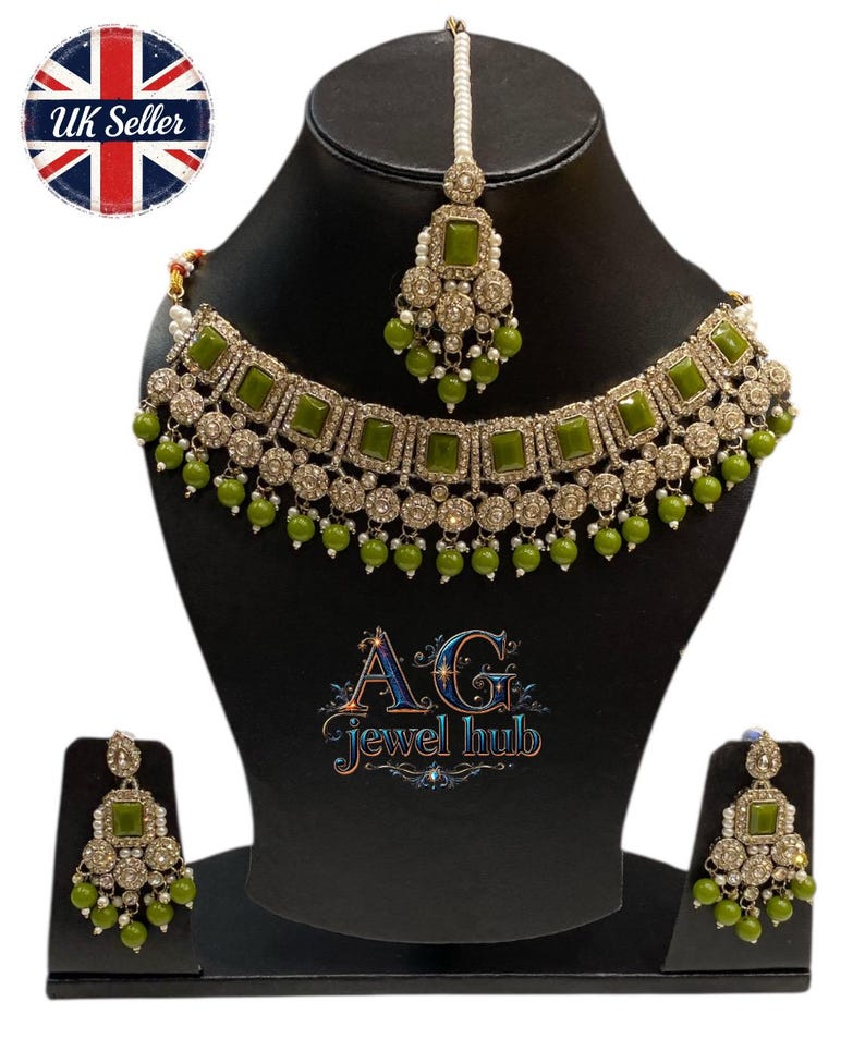 May include: An olive green and silver-toned jewelry set, including a necklace, earrings, and a headpiece. The necklace and headpiece feature square green stones and dangling green beads. The earrings mirror the design. The jewelry is displayed on a black mannequin.