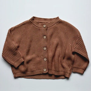 May include: A brown, knitted cardigan sweater with a round neckline and long sleeves. The sweater has a button-up front with five wooden buttons. The knitwear is a solid color and appears to be made of a soft, textured material.