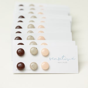 May include: A set of three pairs of small, round, hypoallergenic earrings in brown, beige, and light pink. The earrings are displayed on a white card with the text "sensitive ears studs" and the brand name "sensitive".