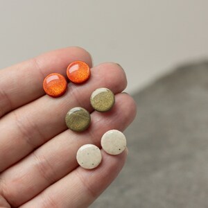 May include: Six small round ceramic earrings in three colors: orange, green, and white. The earrings are displayed on a hand.